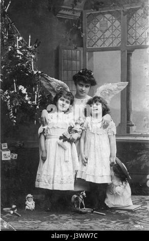 A mother dressed as an Angel, with two girls, probably her daughters, c. 1910. They stand by a Christmas tree decorated with lighted candles, coiled paper strips, and strings. The girls hold new dolls, and also visible are a toy horse, toy blocks and toy badminton (?) rackets. 1910.   To see my other Christmas-related vintage images, Search:  Prestor  vintage  holiday Stock Photo