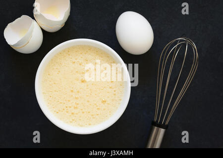 Whisked whole eggs (whites and yolks together) in bowl, with whisk and egg shells on the side, photographed overhead on slate with natural light Stock Photo