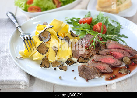 Fried beef tenderloin with Italian pasta and slices of fresh black autumn truffles, served with a mixed side salad and baguette Stock Photo