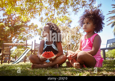 Two young sisters, sitting on grass, eating watermelon Stock Photo