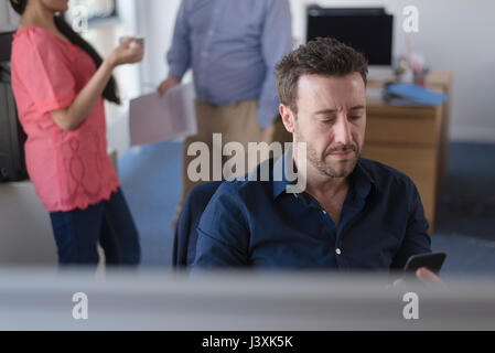 Male office worker checking mobile phone at desk, colleagues on coffee break in background Stock Photo