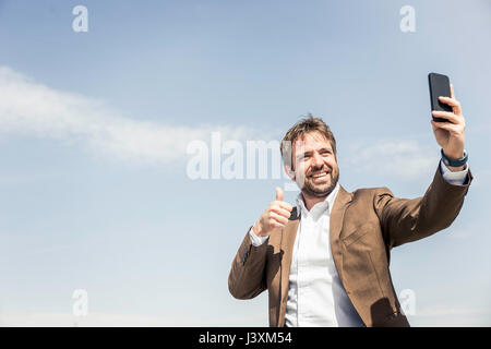 Businessman giving thumbs up for smartphone selfie against blue sky Stock Photo