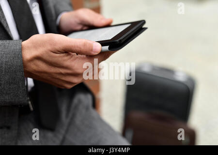 closeup of a young caucasian businessman in a gray suit using a tablet outdoors, next to his luggage Stock Photo
