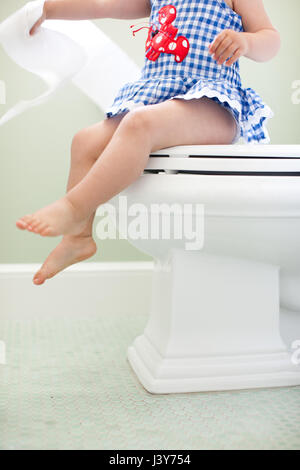 Neck down view of female toddler sitting on toilet seat pulling toilet paper Stock Photo