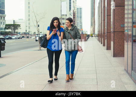 Two female friends walking along street, looking at smartphone