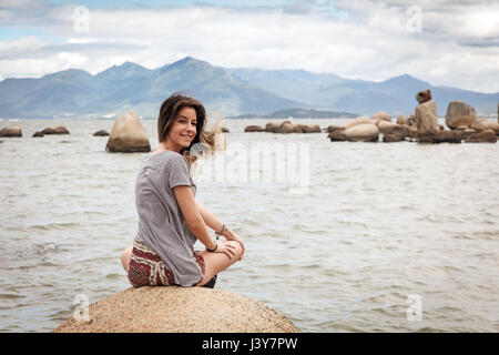 Rear view of teenage girl sitting on rocks looking over shoulder at camera smiling Stock Photo