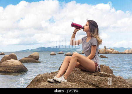 Teenage girl sitting on rocks drinking from water bottle Stock Photo
