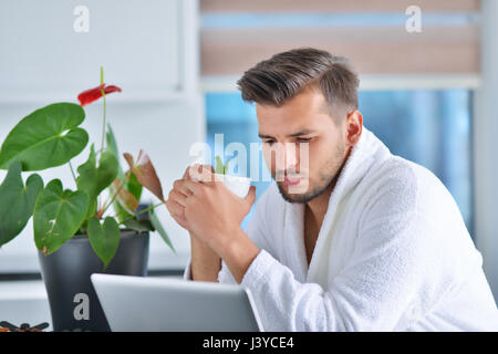Handsome young man drinking coffee while working with laptop in kitchen Stock Photo