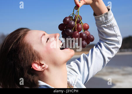 Beautiful young girl eating red grapes outdoor on blue sky background Stock Photo