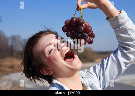 Beautiful young girl eating red grapes outdoor on blue sky background Stock Photo