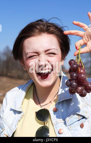 Beautiful young girl eating red grapes outdoor on blue sky background Stock Photo