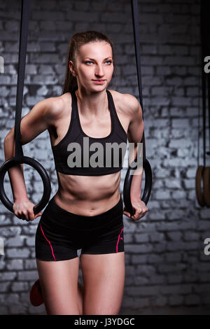 Beautiful female sporty woman using rings equipment in cross fit gym against brick wall. Stock Photo