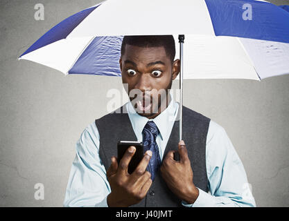 Closeup portrait shocked, surprised business man, corporate executive reading bad, breaking news on smart phone holding umbrella protected from rain i Stock Photo