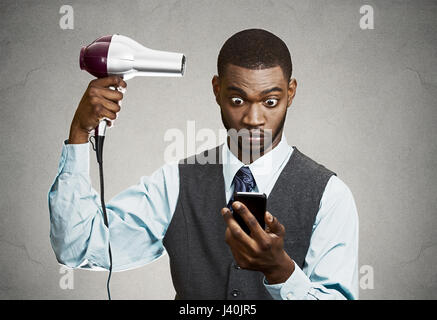 Closeup portrait worried, shocked business man, deal maker reading bad news on smart, mobile phone holding hairdryer isolated black grey background. H Stock Photo