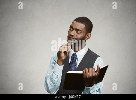 Closeup portrait confused, unhappy serious man holding, reading  book, having many questions, thinking, isolated black background. Human face expressi Stock Photo