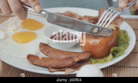 Woman cut tomato in full breakfast with fried eggs, sausages, bacon, potato in big white plate. Female eat in restaurant. 4K Stock Photo