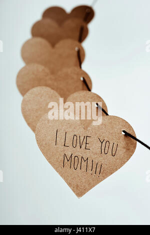 closeup of some brown cardboard hearts strung on a string and the text I love you mom written in the first one, against an off-white background Stock Photo