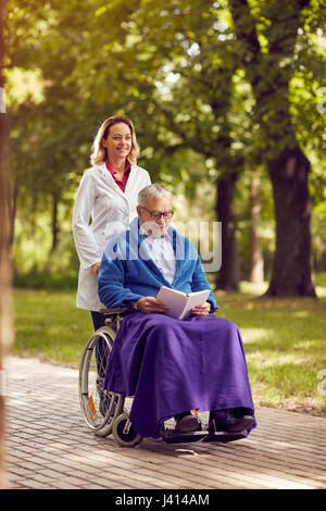 smiling nurse pushing elderly man in wheelchair while reading book in the park Stock Photo