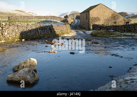 Icy ford across stream near Malham, Yorkshire Dales. Dry stone walls and barn. Road to snow covered hills. Stock Photo