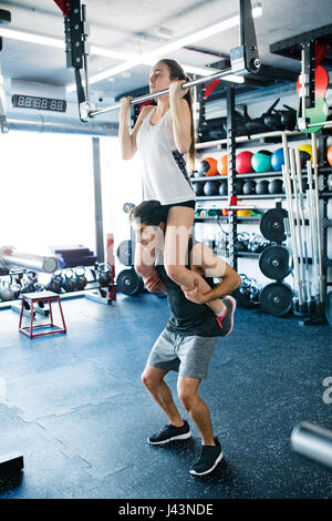 Woman in gym doing pull ups, man giving her piggyback. Stock Photo