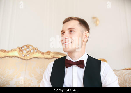 Groom sitting on the sofa waiting for the bride on his wedding day. at wedding tuxedo smiling and waiting for bride.Elegant man in black costume and b Stock Photo