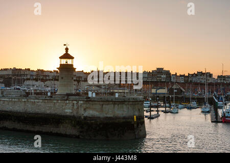 Ramsgate Royal Harbour. Lighthouse at end of jetty with boats moored in the harbour and behind that Ramsgate seafront town with the sun setting over. Clear blue sky other then couple of small white clouds. Stock Photo
