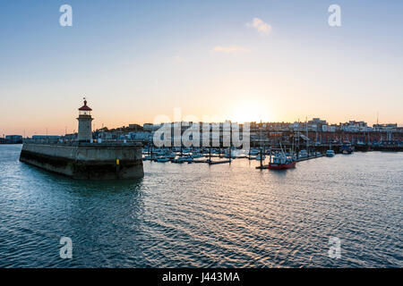 Ramsgate Royal Harbour. Lighthouse at end of jetty with boats moored in the harbour and behind that Ramsgate seafront town with the sun setting over. Clear blue sky other then couple of small white clouds. Stock Photo