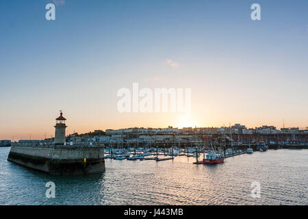 Ramsgate Royal Harbour. Lighthouse at end of jetty with boats moored in the harbour and behind that Ramsgate seafront town with the sun setting over. Clear blue sky other then couple of small white clouds. Stock Photo