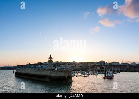 Ramsgate Royal Harbour. Lighthouse at end of jetty with boats moored in the harbour and behind that Ramsgate seafront town with the sun setting over. Clear blue sky other then couple of small white clouds. Stock Photo