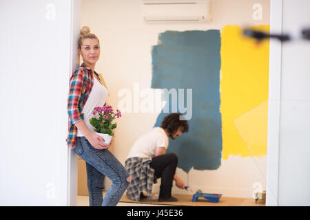 Happy couple doing home renovations, the man is painting the room and the woman hold the pot with flowers Stock Photo