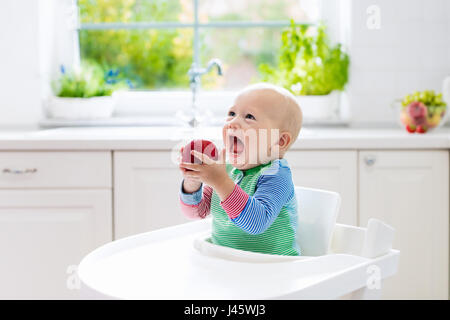 Baby eating fruit. Little boy biting apple sitting in white high chair in sunny kitchen with window and sink. Healthy nutrition for kids. Solid food f Stock Photo