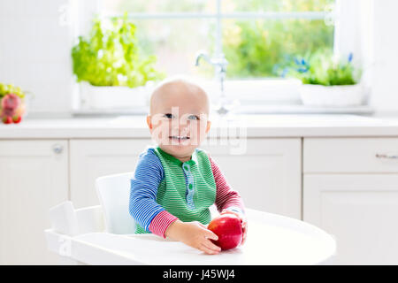 Baby eating fruit. Little boy biting apple sitting in white high chair in sunny kitchen with window and sink. Healthy nutrition for kids. Solid food f Stock Photo