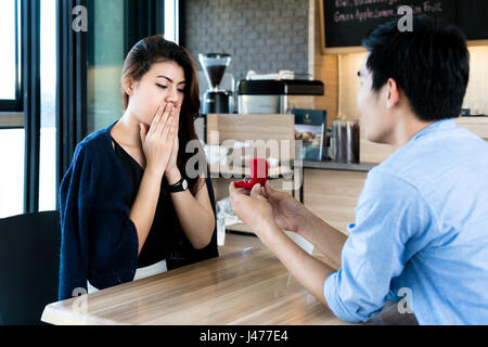 Asian man showing an engagement ring diamond to his amazed girlfriend in a restaurant with a warmth sunset outdoor in the background. Proposal concept