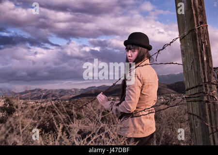 Native American Indian woman in a black hat stands at a barbwire fence at the edge of a indian reservation with a dramatic cloudy sky behind her Stock Photo