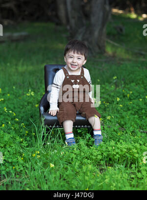 Cute laughing little boy wearing brown corduroy overalls sitting in a blue leather chair in a green meadow Stock Photo