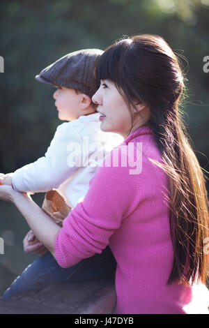 Young and beautiful Asian mother with long black hair wearing a pink cashmere sweater holding her son in a brown newsboy cap as the sun light her hair Stock Photo