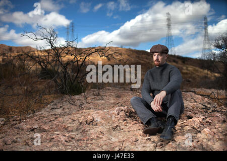 Sad and lonely man with a red goatee wearing a grey wool turtleneck sweater and brown newsboy cap sitting on a rocky mountain peak with white clouds Stock Photo