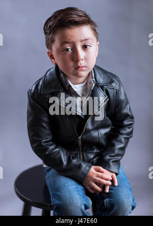 Punk rock and roll little boy with rockabilly greaser haircut wearing a black leather motorcycle jacket and blue denim jeans Stock Photo