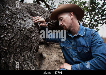 Portrait of a sad cowboy with a red goatee wearing a blue denim chambray cowboy shirt and brown stetson cowboy hat leaning on a oak tree Stock Photo