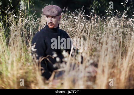 Man with red goatee wearing black turtleneck fisherman's sweater and brown newsboy cap sitting in a golden field of wheat at sunset Stock Photo