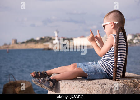 little boy in vest and shorts sitting on breakwater on background of sea and shore Stock Photo