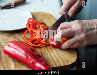 Woman is chopping red bell pepper with knife on a wooden board in the kitchen. Female hands cutting vegetables for salad. Healthy natural food with vi Stock Photo