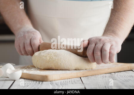 chief man cooks pelmeni or dumplings or ravioli in special mold. Flatten dough with wooden rolling pin on rustic table, front view Stock Photo