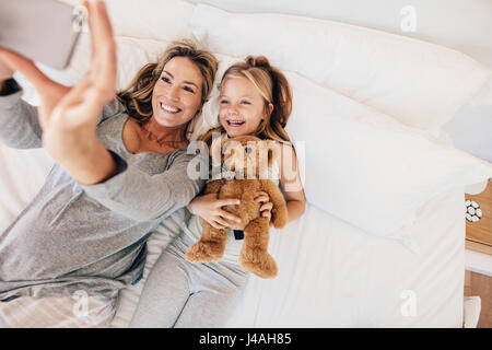 Top view shot of happy young family taking selfie with smartphone in the bed. Mother and daughter lying on bed taking self portraits in bedroom.