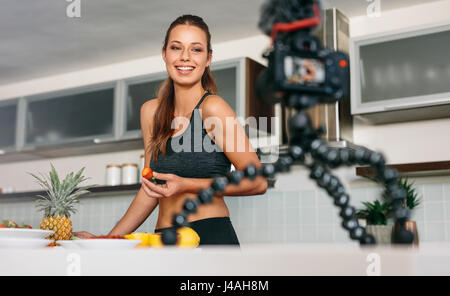 Young lady holding a strawberry at the kitchen table facing the camera. Smiling woman recording content for her vlog in kitchen. Stock Photo