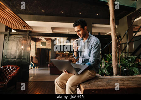 Shot of young man sitting relaxed looking at laptop and having coffee. Male executive using laptop during coffee break. Stock Photo