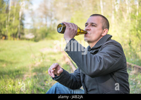 Adult man sitting on rails drinking beer and smoking a cigarette, close-up, with blurred background Stock Photo
