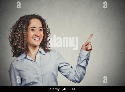 Closeup portrait happy confident young smiling woman gesturing, presenting pointing finger copy space at right isolated grey wall background. Positive Stock Photo