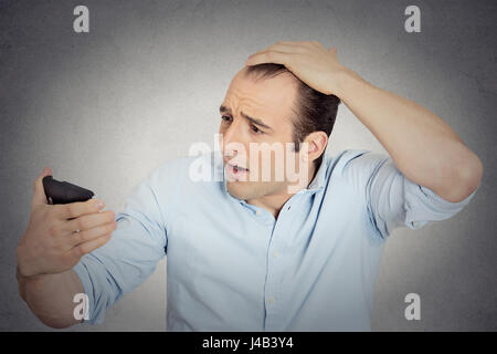 Closeup portrait shocked man feeling head, surprised he is losing hair, receding hairline or seeing bad news on cell phone isolated grey wall backgrou Stock Photo
