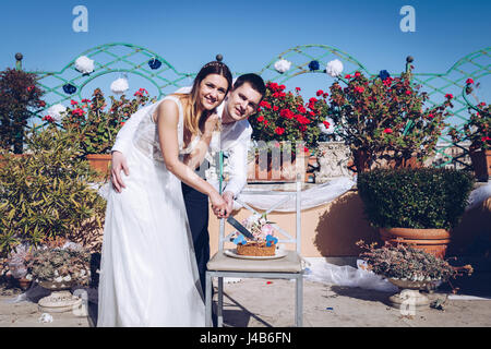 Beauty bride and handsome groom are cutting a wedding cake. Couple on the terrace with Rome cityscape in the background. Beautiful model girl in white Stock Photo